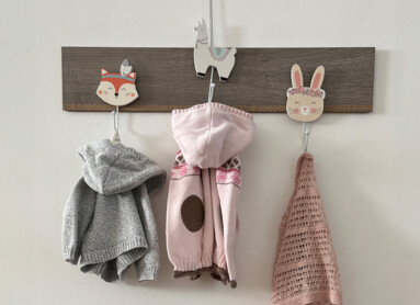 Kinderkleidung an Wandhaken über einer Holzkommode mit Kinderschuhen und grauem Fellkissen.