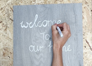Person schreibt "welcome to our home" auf graue Oberfläche.