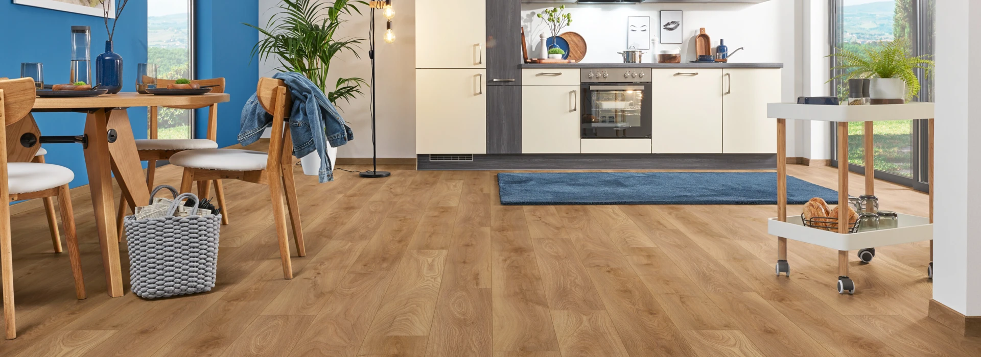 Moderner Küchen- und Essbereich mit hellem Laminatboden der Serie 'Aquaprotect Smart Silent' und blauer Wand, Holzmöbeln und großen Fenstern.