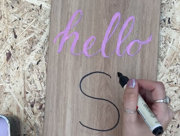 Eine Hand schreibt "S" auf ein Holzbrett mit "hello" in rosa Schrift.