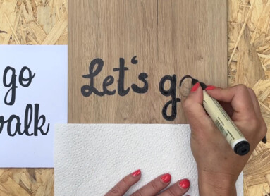 Eine Hand schreibt "Let's go" auf Laminatboden mit schwarzem Stift. Im Hintergrund liegt eine Schablone und ein Farnblatt.