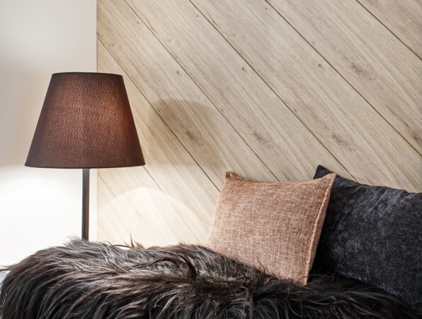 Gemütliche Ecke mit schwarzem Fellüberwurf, braunem und schwarzem Kissen auf Holzbank, daneben steht eine braune Lampe vor einem Wandpaneel der Serie 'decoration'