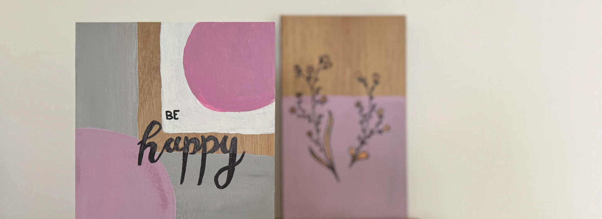 Zwei bemalte Holztafeln auf einem Tisch, eine mit der Aufschrift "Be Happy" in Rosa und Grau.