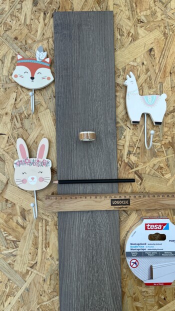 Dekorative Haken, ein Lineal, Bleistift, doppelseitiges Klebeband und ein Laminatstück auf Spanplatte angeordnet.