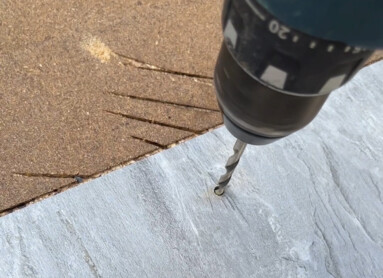 Eine Hand hält eine Bohrmaschine, die ein Loch in eine graue Platte bohrt.