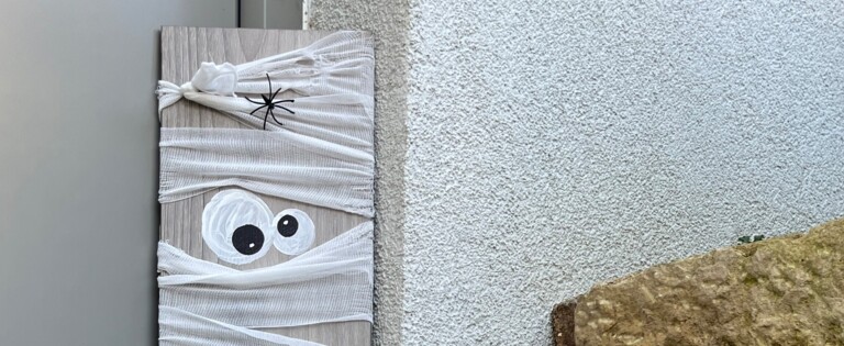 Eine DIY-Halloween-Dekoration mit Mumienfiguren aus Holz und Bändern, dekoriert mit Spinnen und Augen, neben einer Laterne im Freien.