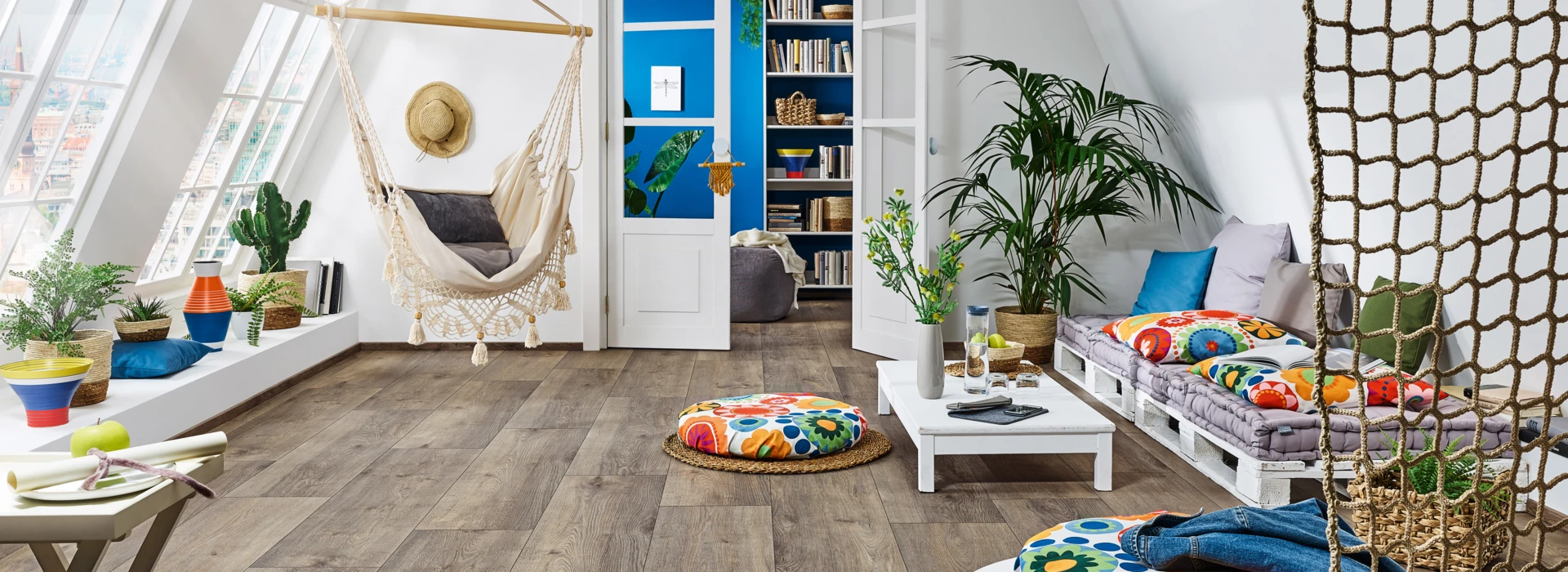 Bright room with laminate flooring, featuring a hammock, colourful cushions, plants, and a bookshelf, creating a cosy and inviting atmosphere.