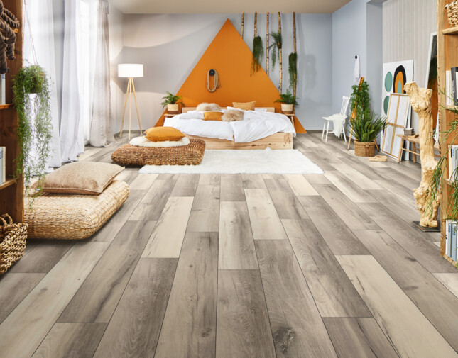 Offenes Schlafzimmer mit hellem Laminatboden, Pflanzen und natürlichen Materialien, im Hintergrund ein großes Bett mit orangenem Akzent.