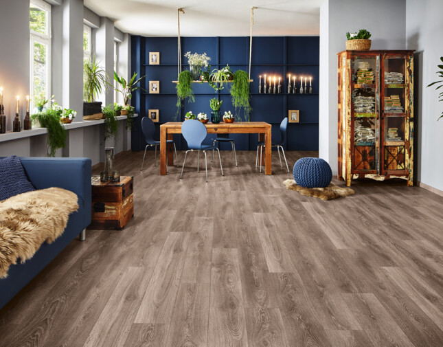 Modern eingerichtetes Wohnzimmer mit dunkelbraunem Laminatboden, blauer Couch, Holztisch und Pflanzen.