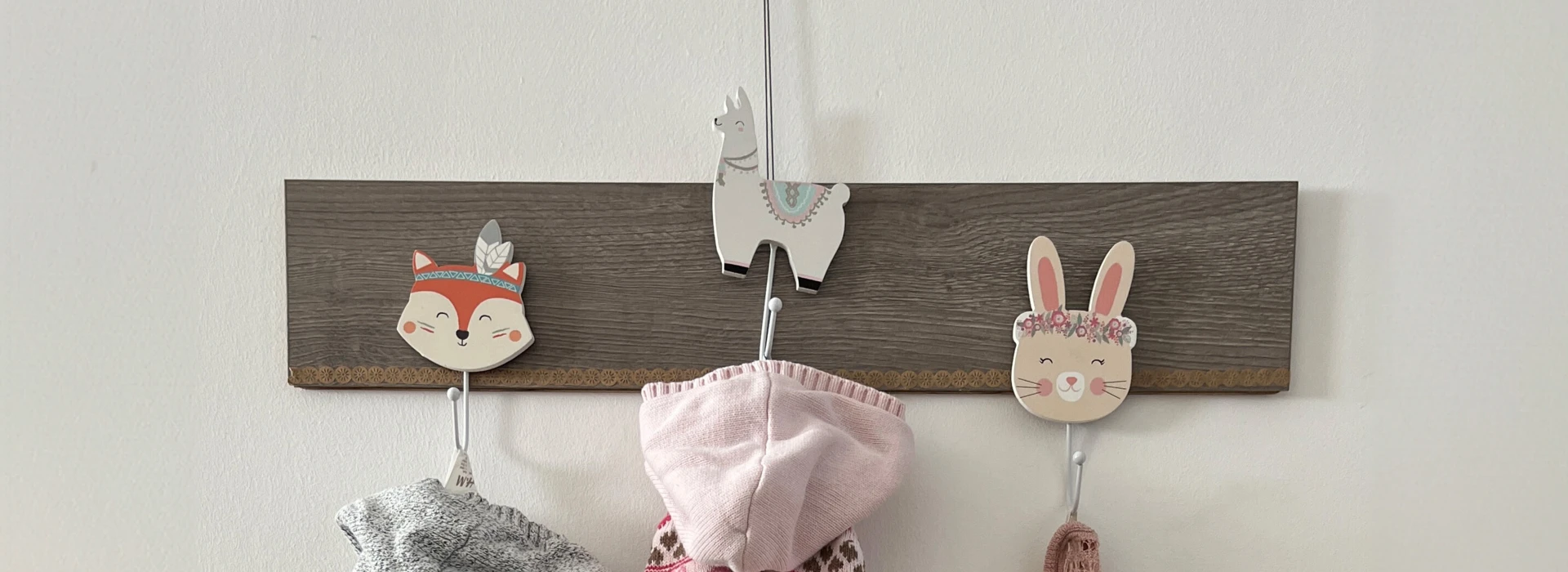 Kinderkleidung an Wandhaken über einer Holzkommode mit Kinderschuhen und grauem Fellkissen.