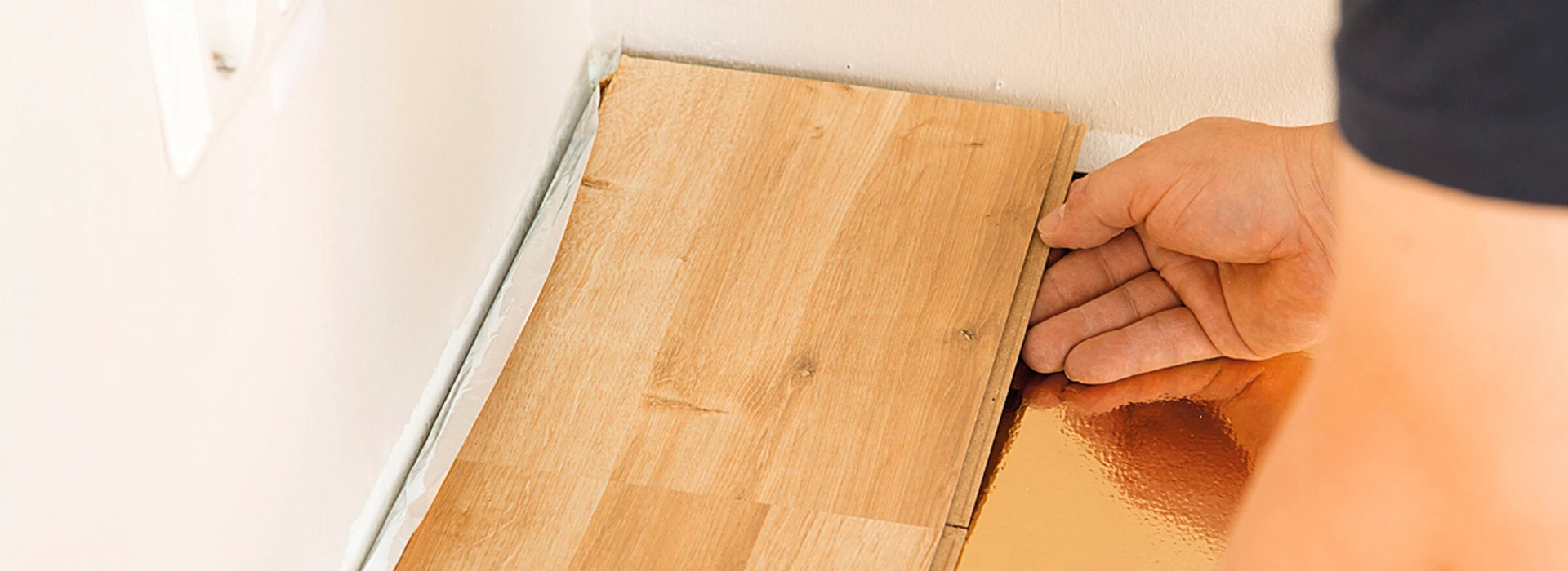 Ein Handwerker verlegt Laminatboden entlang einer Wand mit Steckdosen im Hintergrund.