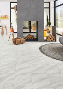 Wohnzimmer mit hellem Laminatboden, grauem Sofa, Kamin und großen Fenstern. Im Hintergrund eine offene Küche mit Dekoration.