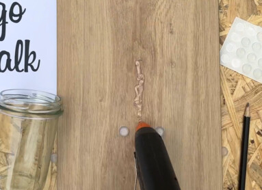 Eine Hand benutzt eine Heißklebepistole auf einer Holzplatte mit der Aufschrift "Let's go for a walk", umgeben von Bastelmaterialien.