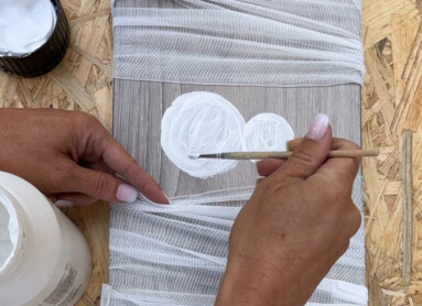 Eine Hand malt weiße Kreise auf eine mit Stoffstreifen umwickelte Holzplatte.