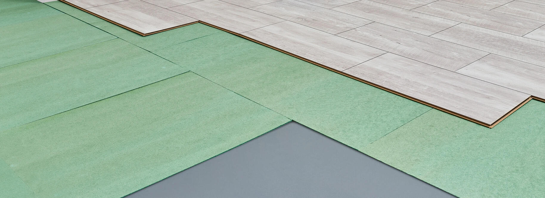 Laminatbodenverlegung mit Werkzeug, Leiter und grünem Unterboden in einem Raum mit großem Fenster.
