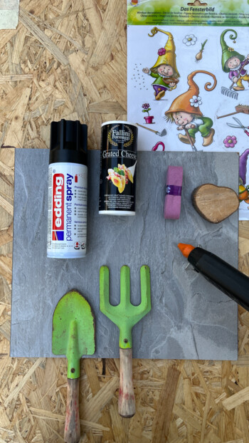 Verschiedene Bastelmaterialien und Werkzeuge, darunter eine Sprühdose, eine Dose von geriebenem Käse, Gartenwerkzeuge und Aufkleber auf Laminatboden.