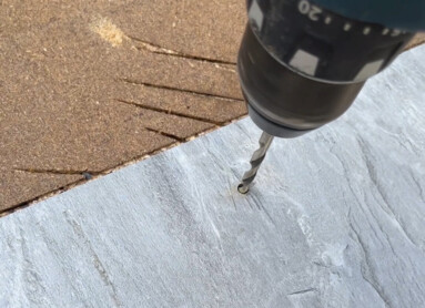 Eine Hand hält eine Bohrmaschine, die ein Loch in eine graue Platte bohrt.