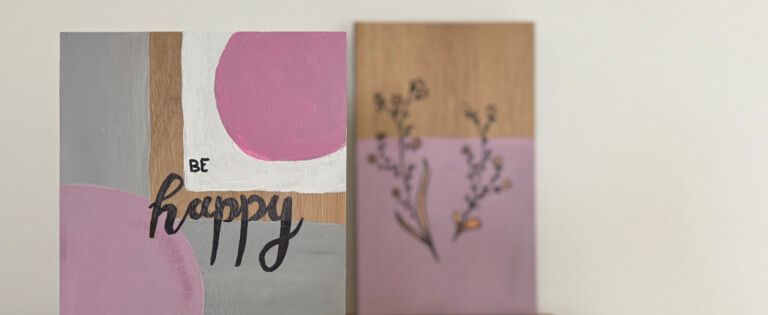 Zwei bemalte Holztafeln auf einem Tisch, eine mit der Aufschrift "Be Happy" in Rosa und Grau.