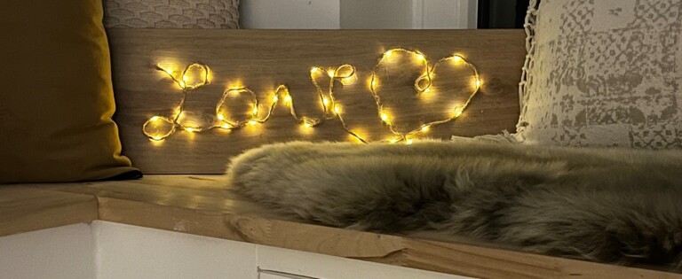 Gemütliche Ecke mit Sitzkissen und Lichterkette, die "Love" auf einem Holzbrett formen.