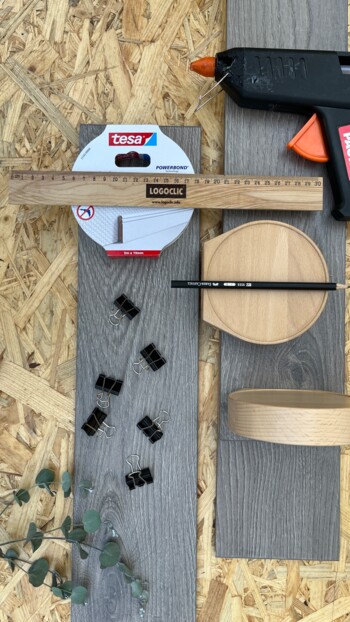 Werkzeuge und Materialien für die DIY Postkartenwand, darunter Laminatboden, Klebepistole, Lineal, Klebeband, Klemmen, Bleistift und Pflanzenzweig.